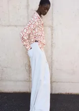 Camisa de corte recto con cuello camisero, manga larga con puños abotonados y cierre frontal de botones ocultos bajo tapeta. Presenta un diseño con estampado abstracto en tonos rojizos sobre fondo claro.