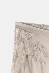Pantalón de tiro alto confeccionado en tejido satinado color beige. Presenta pernera recta, cintura elástica y aberturas laterales ajustables con lazadas. Incluye aplicaciones de encaje a tono en los laterales.