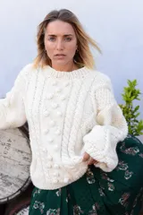 Sweater artesanal de tejido de punto de lana merino color marfil, de calce amplio y relajado, con cuello redondo y diseño frontal en relieve con trenzas y bodoques. Presenta mangas largas abullonadas con paneles texturizados y puños acanalados.