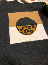 Campera de textura tipo bouclé en color negro, con un aplique central de diseño geométrico que combina bloques de color en beige, blanco y un círculo con estampado animal print.