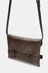 Bolso formato bandolera de material sintético con acabado metálico plateado. Presenta un diseño de solapa frontal con un cinturón decorativo que se cierra con una hebilla metálica en forma de corazón. Incluye un asa bandolera fina de cadena y un bolsillo interior con cierre.