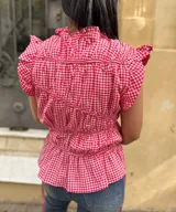 Blusa sin mangas con estampado vichy en rojo y blanco, cuello redondo fruncido y volados en los hombros. Presenta frunces horizontales en el frente, lazos laterales regulables y bajo con ligero vuelo.