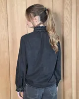 Camisa de denim negro con volados en el cuello alto y en la botonera frontal, manga larga con puños fruncidos y cierre de botones metálicos dorados.