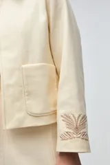 Chaqueta de lana fina sin teñir, con cuello clásico y bolsillos frontales. Presenta detalles bordados en el cuello y en los puños de las mangas. Es una prenda forrada con diseño exclusivo.
