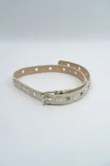 Cinturón de cuero sintético color beige, con hebilla metálica plateada y diseño decorado con ojales metálicos y apliques circulares grabados a lo largo de toda la tira.