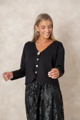 Cardigan de punto fino color negro con escote en V, manga larga y cierre frontal con botones dorados labrados, destacando un botón joya superior en forma de flor.