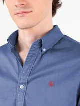 Camisa azul de hombre confeccionada en 100% algodón twill, tintada en prenda. Cuenta con corte regular fit, cuello button-down, cierre frontal de botones, manga larga con puño simple y pequeño logo rojo bordado en el pecho.