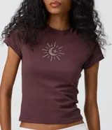 Remera baby tee de algodón color marrón oscuro, con cuello redondo, manga corta y calce ajustado. Presenta un aplique frontal de pedrería brillante con diseño místico de sol, luna y estrella.