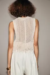 Chaleco de punto calado color crudo, de cuello redondo y sin mangas. El bajo está terminado con flecos.