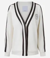 Cardigan deportivo de corte oversized, color blanco con detalles en marrón oscuro. Presenta escote en V, cierre frontal con botones a presión, mangas largas con franjas laterales y puños acanalados. Incluye un parche circular con letras en el pecho.