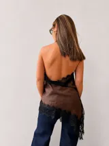Blusa de inspiración lencera color marrón, con escote buche y detalles de encaje negro en los bordes. Presenta un diseño asimétrico con ruedo en pico y espalda descubierta.