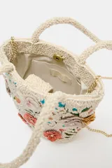 Bolso de mano de formato tipo bucket en color crudo, completamente revestido con abalorios y bordado con motivos florales multicolores en tonos rosados, verdes y celestes. Cuenta con doble asa corta de mano y cierre con botón imantado.