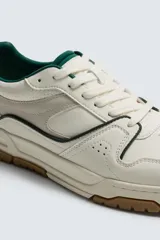 Zapatilla deportiva de estilo retro con diseño de paneles en tonos crema y blanco, detalles en verde oscuro y suela de goma color caramelo.
