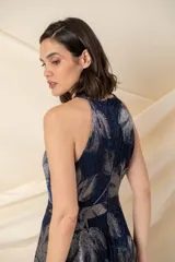 Vestido largo de fiesta en gasa plisada color azul con estampado plateado de hojas. Tiene falda amplia, cruzada y abierta, con bolsillos laterales. El escote es halter y tiene un cinto fino a la altura de la cintura.