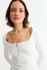 Remera de manga larga, color blanco, con tejido acanalado y escote cuadrado. Presenta un cierre metálico dorado en el centro del escote.