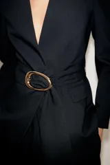 Blazer negro de cuello sin solapa y manga larga con hombreras. Presenta un cinturón integrado con hebilla dorada, bolsillos delanteros con solapa y cierre frontal con un botón.