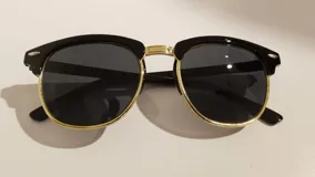Lentes de sol estilo Clubmaster con armazón superior negro, inferior dorado y lentes grises.