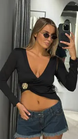 Top negro de manga larga con escote en V profundo y diseño cruzado. Presenta un detalle metálico circular dorado en el lateral de la cintura y un corte asimétrico que deja parte del abdomen al descubierto.
