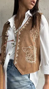 Chaleco de gamuza color camel con bordados en hilo blanco y detalles de tachas metálicas. Presenta un diseño abierto con terminaciones redondeadas en el frente.