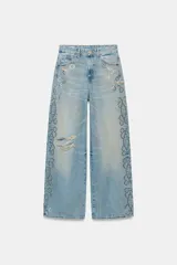 Jeans de tiro bajo y corte wide leg, confeccionados en denim de color celeste claro con lavado desgastado. Presentan detalles de roturas y aplicación de tachas metálicas en los laterales y bolsillos delanteros. Cierre frontal con cremallera y botón.