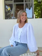 Blusa blanca de algodón con textura arrugada, cuello redondo, mangas largas abullonadas con puños elásticos y cierre frontal con botones y volantes.