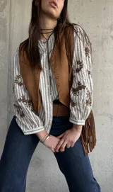Chaleco de gamuza color camel forrado, con ribetes decorados con tachas metálicas y tiras colgantes con flecos en el lateral.