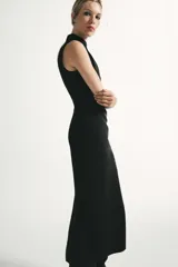 Vestido midi negro con cuello subido y diseño sin mangas (sisa). Presenta un elegante detalle de tejido drapeado en el lateral y cierre trasero con cremallera oculta.