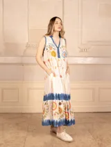 Vestido largo color crema con estampado de flores y corazones en tonos azules, naranjas y amarillos. Tiene cuello en V con ribete azul y corte imperio con falda escalonada.