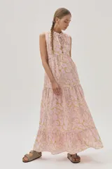 Vestido largo de algodón color rosa con estampado floral en tonos beige y dorado. Tiene cuello alto con lazo ajustable, corte imperio, falda escalonada con volados y diseño sin mangas.