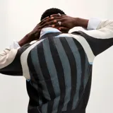 Sweater de tejido punto con diseño inspirado en camisetas de fútbol. Cuerpo principal en gris oscuro y negro con franjas verticales celestes. Presenta un gran logo "Nude Project" en blanco en el frente, un escudo bordado con una rosa roja en el pecho y mangas raglán en gris claro.