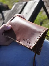 Billetera y tabaquera de cuero marrón con costuras decorativas artesanales en el contorno y cierre con botón a presión. Cuenta con compartimentos interiores para accesorios y tarjetas.