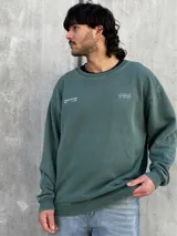 Buzo verde de algodón con cuello redondo, mangas largas y puños acanalados. Presenta un pequeño estampado en el frente y un logo geométrico en el pecho. Corte holgado.