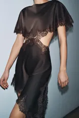 Vestido midi negro de tejido satinado con cuello redondo y manga capa. Presenta detalles de cut-out laterales adornados con encaje a tono. El bajo es asimétrico y tiene cierre en la espalda.