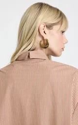 Camisa de corte recto y diseño oversize, con estampado de rayas verticales en tonos marrón y blanco. Presenta cuello clásico estructurado, mangas largas con puños anchos, hombros caídos y bajo asimétrico con abertura lateral. Cierre frontal mediante botones.