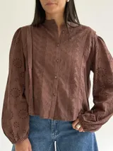 Blusa de brodery color blanco, con cuello redondo, mangas largas abullonadas con puños elásticos y cierre frontal con botones forrados. Presenta detalles de puntillas verticales en el frente.