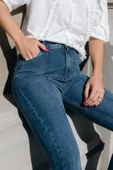 Jean de denim azul de corte acampanado, con costuras frontales en relieve tipo alforza y terminación desmechada en el bajo.