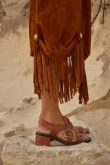 Sandalias de cuero marrón con tiras anchas cruzadas y hebilla metálica decorativa en forma de estribo. Poseen taco cuadrado de altura media y suela de goma negra.