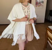 Ruana tejida color crudo con flecos en los bordes. Diseño abierto y corte rectangular.