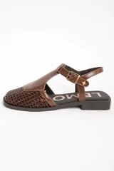 Sandalias de cuero marrón, con puntera cerrada de diseño trenzado, empeine liso y tira ajustable con hebilla en el talón. Cuenta con suela plana y baja.