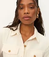 Vestido midi color crema, tipo camisero, con cuello, manga corta y cierre frontal de botones. Presenta bolsillos utilitarios en el pecho y la cadera, y un cinturón ajustable con hebilla redonda de madera. Confeccionado en sarga de algodón.