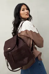 Mochila de símil gamuza color marrón oscuro con textura tipo reptil. Presenta un diseño con solapa frontal, cierre de cordón ajustable, bolsillos laterales y herrajes metálicos dorados.