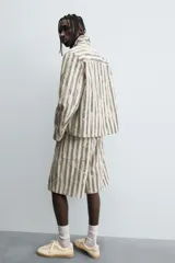 Bermuda de hombre color beige con estampado de rayas verticales azules con efecto desgastado. Corte relaxed fit, con cierre frontal de cremallera y botón, bolsillos delanteros y bolsillos traseros de plastrón.