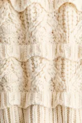 Buzo de punto color beige con textura, cuello redondo acanalado, mangas raglán y detalles de volados horizontales en el frente y las mangas. Presenta puños y ruedo acanalados con terminaciones festoneadas.