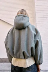 Chaqueta estilo bomber de corte oversize en color marrón oscuro, confeccionada en tejido suave. Presenta capucha con cordones ajustables, cierre frontal con cremallera, mangas largas con puños elásticos y bolsillos delanteros de vivo. El bajo cuenta con detalle de pliegues.
