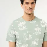Camiseta verde claro con cuello redondo, manga corta y estampado all-over de estilo tropical con palmeras y paisajes costeros.