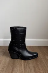 Bota corta de cuero vacuno color negro, con diseño de caña media adornada con pequeñas tachas metálicas distribuidas geométricamente. Presenta un taco cuadrado de altura media y punta ligeramente cuadrada.