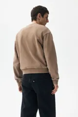 Buzo de tejido pesado de algodón con efecto lavado, color marrón topo. Presenta un diseño estampado en el frente con el texto "RIVERSIDE SOCIAL CLUB" y un escudo con un remo. Tiene cuello redondo, hombros caídos y puños y bajo acanalados.