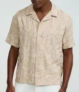 Camisa de poliéster color beige con bordado floral blanco, de corte regular, cuello inglés, manga corta y abotonada en el frente.