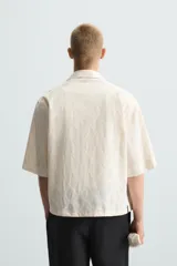 Camisa de corte relajado (relaxed fit) color crema o blanco roto, confeccionada en tejido con mezcla de algodón. Presenta un bordado sutil en relieve con patrón de ondas o líneas diagonales entrecruzadas. Tiene cuello solapa, manga corta y un bolsillo de plastrón en el pecho.