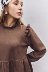 Vestido corto de gamuza color marrón, con diseño de corte evasé, volados en los hombros y mangas largas con puños elásticos fruncidos. Presenta un cuello alto y una falda con un volado en el ruedo.